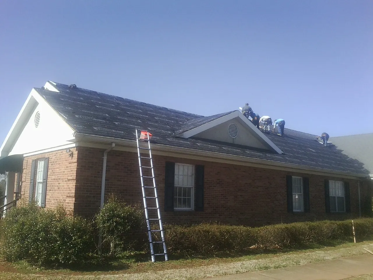 Crew tearing shingles off a brick ranch roof with a ladder set — Emergency Roof Repair work by our crew in Nichols